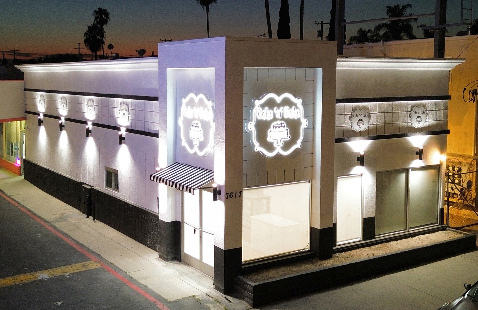 Exterior view of Cake N’ Bake’s bakery at night, showcasing the illuminated logo signs and modern design with a striped awning over the entrance.